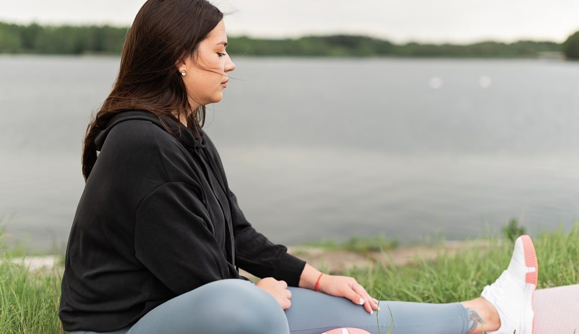 Woman meditating outdoors by a tranquil lakeside, fostering health and wellness.